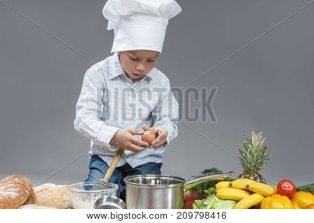 Cuisine Concepts. Portrait of Fascinated Caucasian Little Boy Crashing Fresh Egg In Cooking Hat. Surrounded by Fresh Fruits and Vegetables In Studio. Posing with Smiling Expression. Against Gray. Horizontal Image Composition