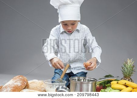 Cuisine Concepts and Ideas.Portrait of Concentrated Caucasian Boy Mixing Fresh Egg in Saucepan. Posing In Cooking Hat Surrounded by Fresh Fruits and Vegetables. Against Gray. Horizontal Image