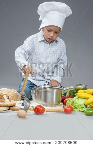 Portrait of Caucasian Boy Working With Kitchen ware In Cooking Hat. Surrounded by Fresh Fruits and Vegetables In Studio. Posing with Smiling Expression. Against Gray.Vertical Image Orientation
