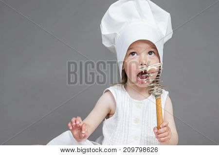 Funny Cooking Concepts. Portrait of Little Caucasian Girl in Cook Uniform Posing with Whisk Covered with Flour. Against Gray. Horizontal Image Composition