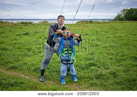 Man teaches boy to control power kite for kitesurfing on green meadow near river.