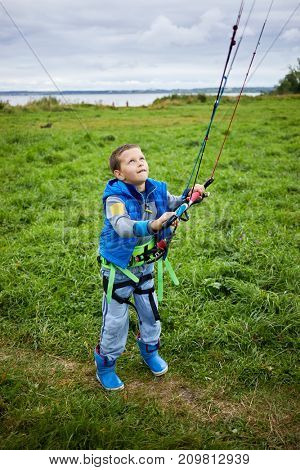 Boy equipped for kitesurfing learns to control power kite on green meadow.