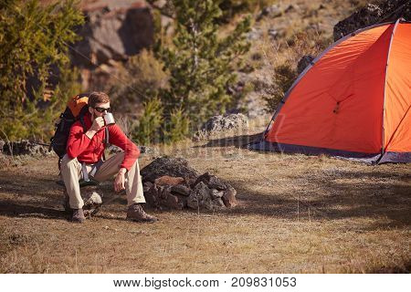 Hicker with beard and sunglasses sitting near orange tent and drink from a cup. Sunny spring daybreak in rocky mountains.