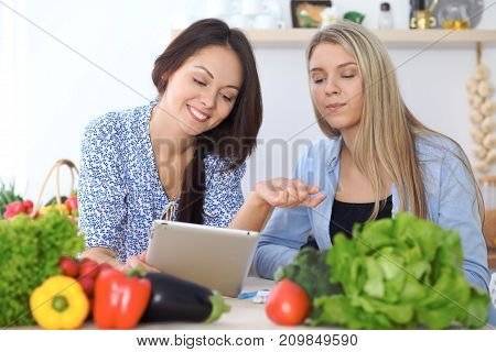 Two young happy women making online shopping for making menu by tablet computer. Friends cooking in the kitchen.