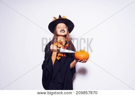 a young sexy gothic girl in the shape of a witch in halloween, in a big black hat, adorned with yellow leaves, holds a pumpkin and a knife in her hand