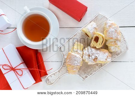 Homemade croissants with fruit jam decorated with powdered sugar with a cup of morning tea on a white wooden background scattered gift white and red boxes tied with a twine. Top view.