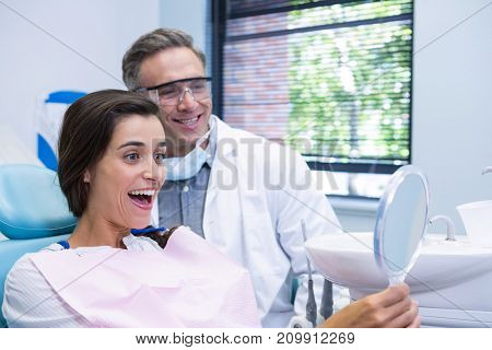 Happy woman looking at mirror by dentist while sitting on chair at medical clinic
