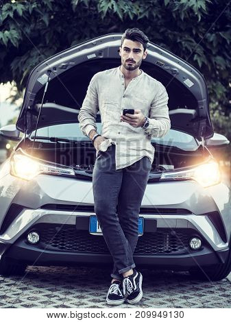 Handsome young man trying to repair a car engine, looking inside open bonnet then calling on cellphone, asking for help