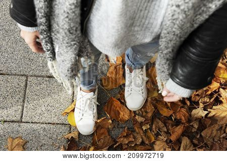Young hipster woman is standing among autumn leaver wearing white sneakers. Top view. Autumn street fashion concept. 