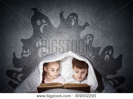 Engrossed Little Kids Reading Book In Bed. Brother And Sister Together Hiding Under Blanket. Fearful
