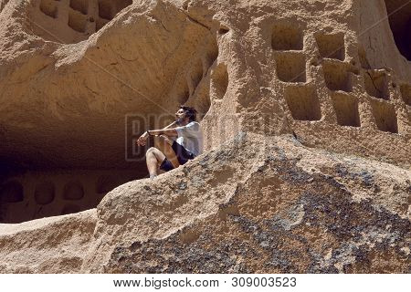 Guy In Shirt And Shorts Sitting On A Rock House With Windows In Uchhisar