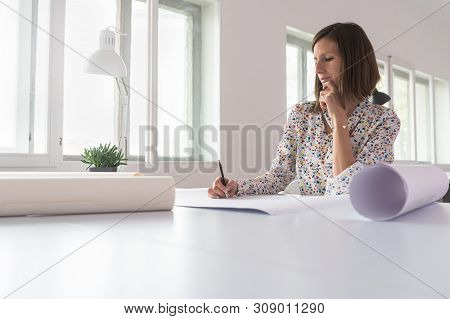 Young Woman Sitting In An Office Working On A Project With Rolled Up Designs Or Blueprints On Her De