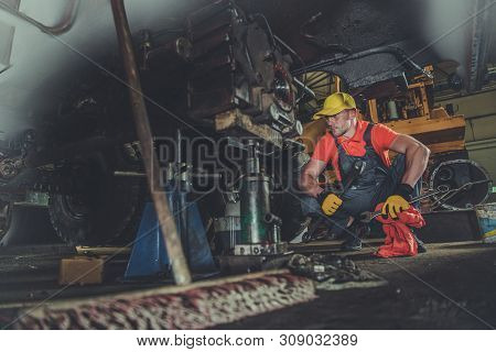 Heavy Equipment Repair Shop. Caucasian Professional Mechanic Fixing Excavator Undercarriage. Industr