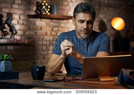 Older white man sitting at desk in dark room at home, working, using tablet