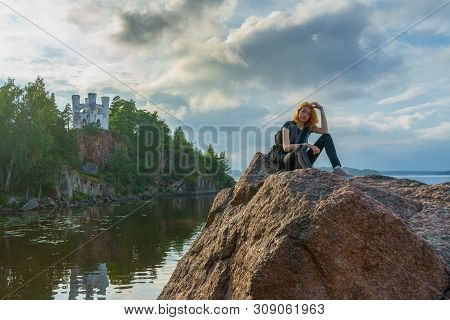 Pretty Young Lady With Red Hair Sitting On Stone Near Bay Shore In Summer Evening. Tourist On The Be