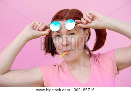 Portrait Of Audacious Young Woman In Pink Shirt With Pigtails Hairstyle, Blue Round Sunglasses And L