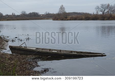 Partially Sunken Boat Near The River Bank, In The Background The River Bank And Houses