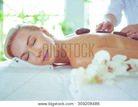 Beautiful Young Woman With Eyes Closed Receiving Hot Stone Massage At Salon Spa.