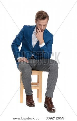 Thoughtful casual man holding his hand on his chin and looking down while wearing a suit and sitting on a chair on white studio background