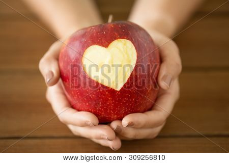 food, valentines day and health concept - close up of hands holding ripe red apple with carved heart shape over wooden table