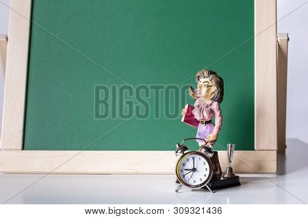 Back To School. Alarm Clock And The Figure Of The Teacher On The Background Of The Blackboard.