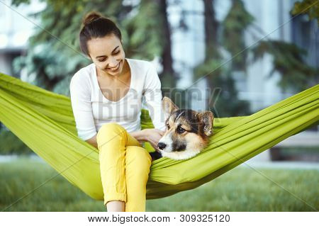 Smiling Young Woman In Green Hammock With Cute Dog Welsh Corgi In A Park Outdoors. Beautiful Happy F