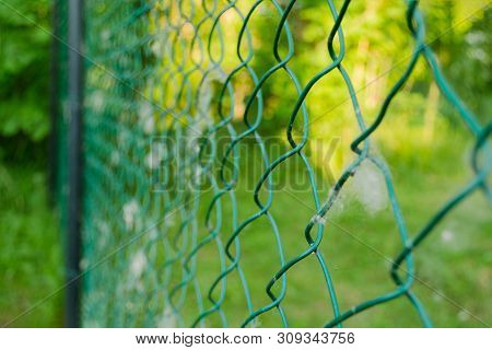 Close Up Of Metal Chain-link In The Garden. Diamond Mesh Wire Fence On Blurred Green Background. Iro