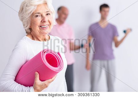 Happy Senior Woman Holding Pink Joga Mat, Men Exercising In Background