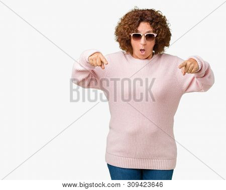 Beautiful middle ager senior woman wearing pink sweater and sunglasses over isolated background Pointing down with fingers showing advertisement, surprised face and open mouth