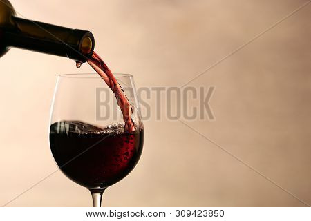 Pouring Red Wine Into Glass From Bottle Against Blurred Beige Background, Closeup. Space For Text