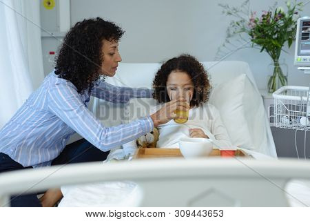 Side view of mixed race mother feeding her son orange juice in bed in the ward at hospital