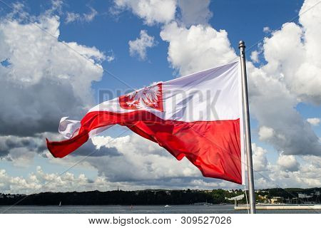 Flag Of Poland. Poland National Flag With Emblem  On Cloudy Blue Sky. Baltic Coast, View From Ship. 