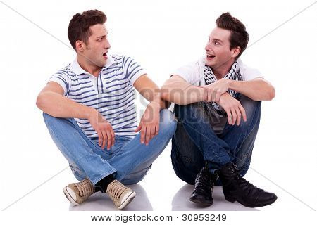 two men friends looking one at the other while sitting next to each other on white background. they both look very surprised