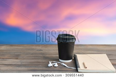 Coffee Cup,ear Phone And Notebook On Wooden Top Withbeautiful Pink Blue Sky  Clouds Background