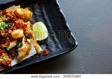 Top View Of Fried Squid With Chili And Lemon In Black Plate On Stone Cutting Board In Restaurant, Re