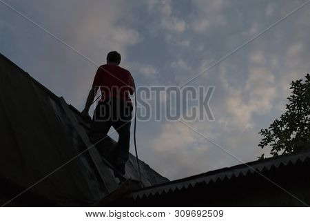 Silhouette Of A Worker With Safety Rope Climbing On The House Roof In Order To Recover Old Tar Paper