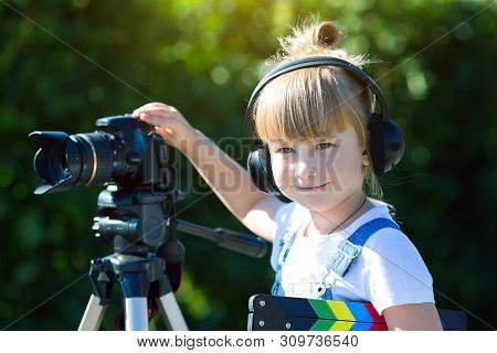 A Little Girl In Headphones With A Tv Numerator In Her Hands Is Standing Next Camera On A Tripod. Po