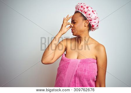 African american woman wearing pink shower towel after bath over isolated background smelling something stinky and disgusting, intolerable smell, holding breath with fingers on nose. Bad smells