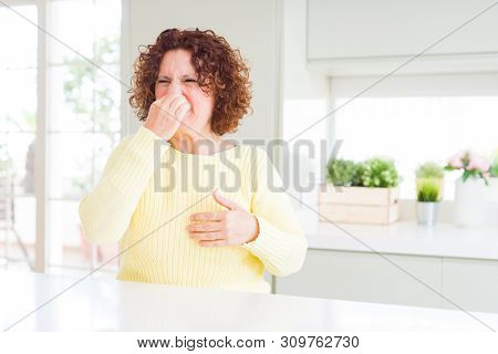Beautiful senior woman wearing yellow sweater smelling something stinky and disgusting, intolerable smell, holding breath with fingers on nose. Bad smells concept.