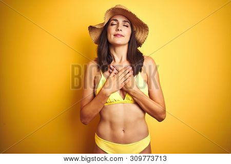 Beautiful woman wearing yellow bikini and summer hat over isolated yellow background smiling with hands on chest with closed eyes and grateful gesture on face. Health concept.