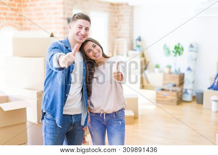 Beautiful young couple moving to a new house doing happy thumbs up gesture with hand. Approving expression looking at the camera with showing success.