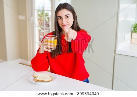 Beautiful young woman eating toasts and orange juice for snack or breakfast pointing with finger to the camera and to you, hand sign, positive and confident gesture from the front