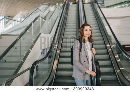 Young Girl At The Airport In Grey Casual Clothes, Standing With Her Suitcase And Backpack Near The E