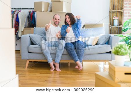 Young couple sitting on the sofa arround cardboard boxes moving to a new house approving doing positive gesture with hand, thumbs up smiling and happy for success. Winner gesture.