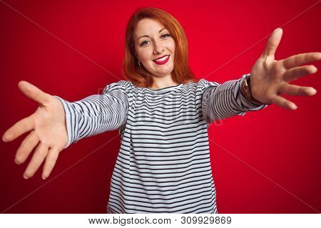 Young redhead woman wearing strapes navy shirt standing over red isolated background looking at the camera smiling with open arms for hug. Cheerful expression embracing happiness.