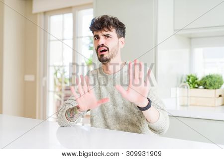 Young man wearing casual sweater sitting on white table afraid and terrified with fear expression stop gesture with hands, shouting in shock. Panic concept.
