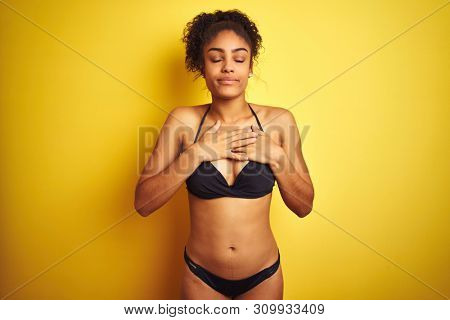 African american woman on vacation wearing bikini standing over isolated yellow background smiling with hands on chest with closed eyes and grateful gesture on face. Health concept.