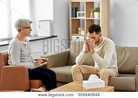 psychology, mental therapy and problem concept - senior woman psychologist taking notes to notebook and sad young man patient at psychotherapy session