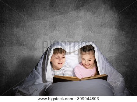 Engrossed Little Girl And Boy Reading Book In Bed After Bedtime. Pretty Kids Together Hiding Under B