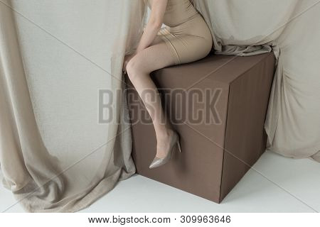 Cropped View Of Adult Model In Dress Sitting On Cube Near Light Curtain